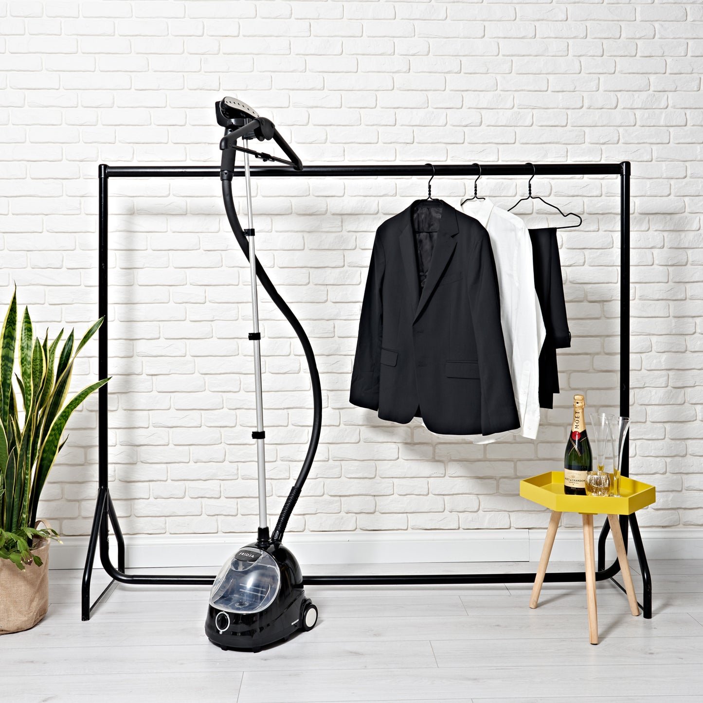 A clothing rack with a white and two black jackets, a Fridja f1000 Clothes Steamer - Refurbished Grade B for refreshing fabrics, a green potted plant, and a yellow table with champagne, glassware, and an ice bucket by a white brick wall.