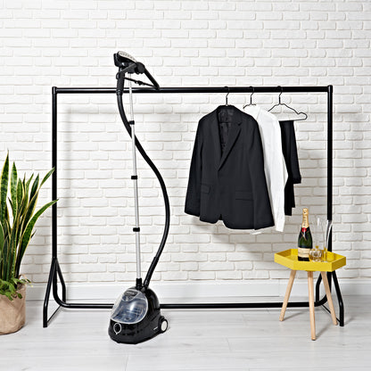A clothing rack with a white and two black jackets, a Fridja f1000 Clothes Steamer - Refurbished Grade B for refreshing fabrics, a green potted plant, and a yellow table with champagne, glassware, and an ice bucket by a white brick wall.