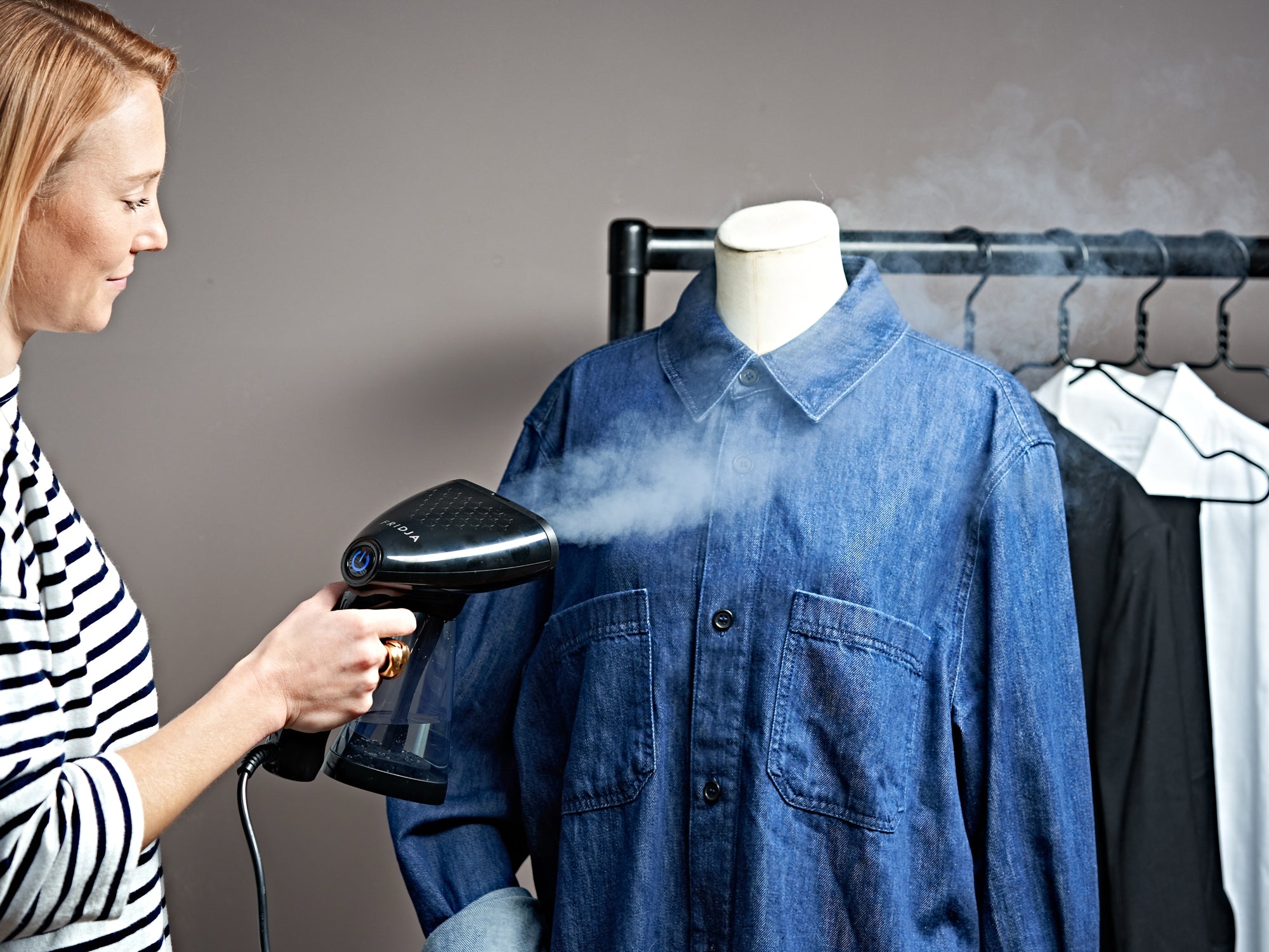 A person uses a handheld steamer to remove wrinkles from a blue denim shirt on a mannequin, with other clothes hanging on a rack in the background.