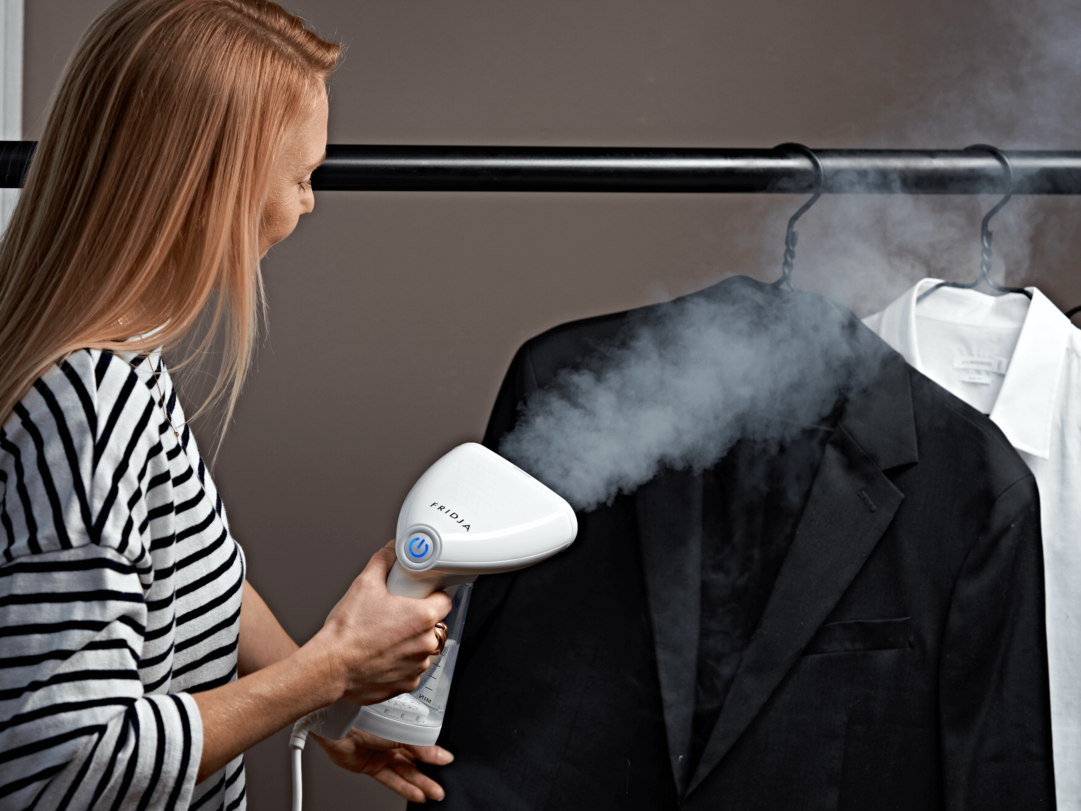 A woman uses a handheld garment steamer to remove wrinkles from a black suit jacket hanging on a clothing rack beside a white shirt. Steam is visible coming from the device.