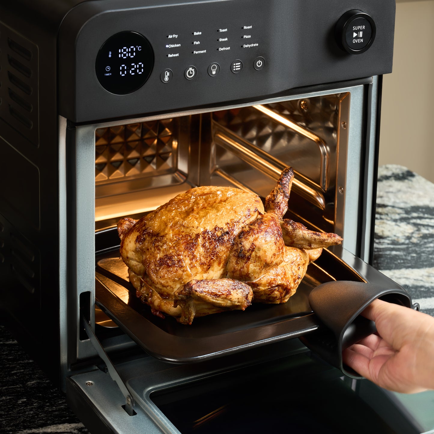 A person removes a cooked whole chicken from the Fridja Super Oven - 15L Digital Air Fryer Oven, featuring a digital display at 180°C and 0:20 left, showcasing the benefits of convection for perfectly roasted meals.