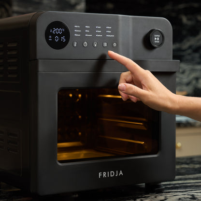 A hand presses a button on the control panel of a black Fridja Super Oven - 15L Digital Air Fryer Oven with digital display and 12 smart presets in a modern kitchen.