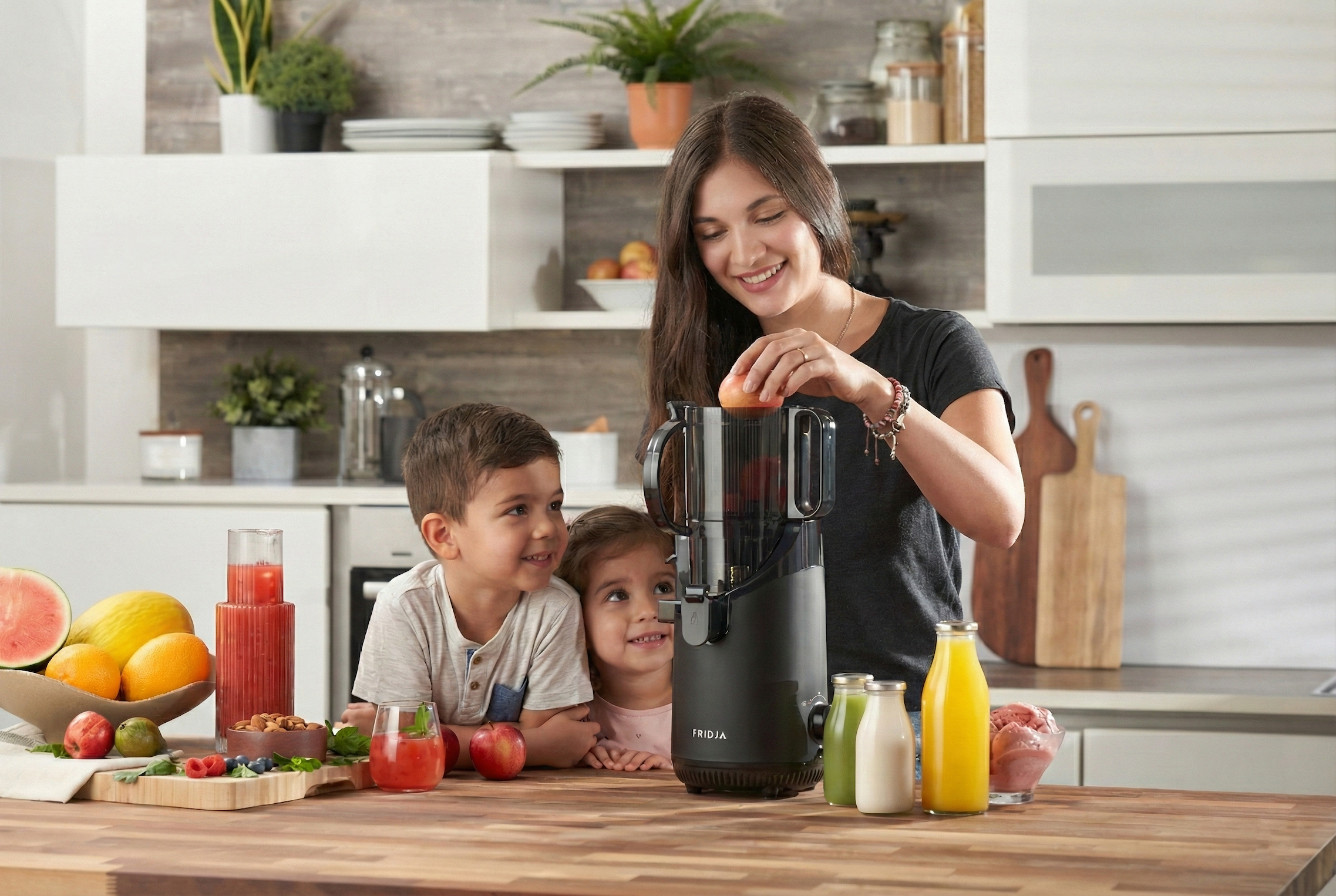A woman uses a juicer to make fresh juice in a modern kitchen while two young children watch and smile. Bottles and glasses of juice, fruit, and vegetables are on the wooden counter.