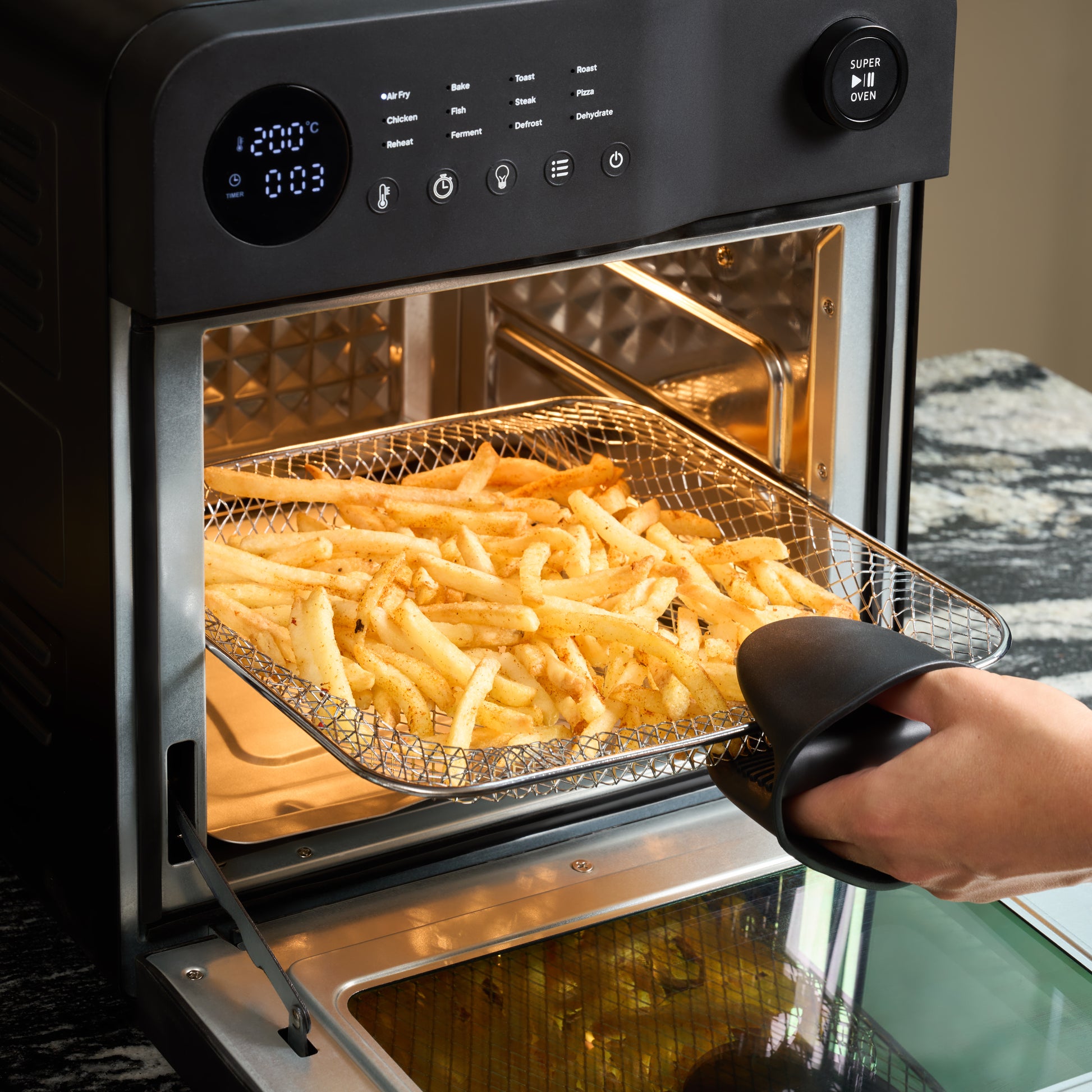 A hand uses an oven mitt to remove a tray of French fries from the Fridja Super Oven - 15L Digital Air Fryer Oven, which displays convection cooking settings on its digital screen on the kitchen countertop.