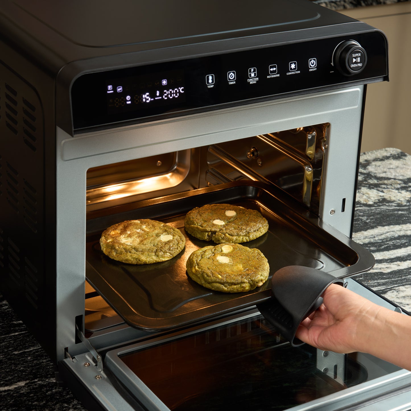 A hand wearing a black oven mitt pulls out a tray with three large cookies from the Fridja f77 Super Oven Pro – 30L Digital Air Fryer Oven, which is PFAS free and displays temperature and cooking settings.