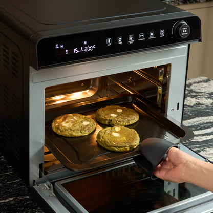A hand wearing a black oven mitt pulls out a tray with three large cookies from the Fridja f77 Super Oven Pro – 30L Digital Air Fryer Oven, which is PFAS free and displays temperature and cooking settings.