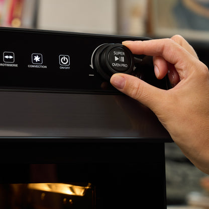 A hand turns the control knob of the Fridja Super Oven Pro - 30L Digital Air Fryer Oven, showing buttons for rotisserie, convection, and on/off on its control panel.