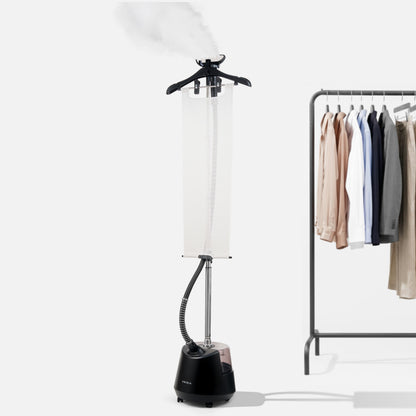 A Fridja f1200 Clothes Steamer Pro emits steam as it stands beside a clothing rack displaying a variety of shirts and jackets on hangers against a plain background.