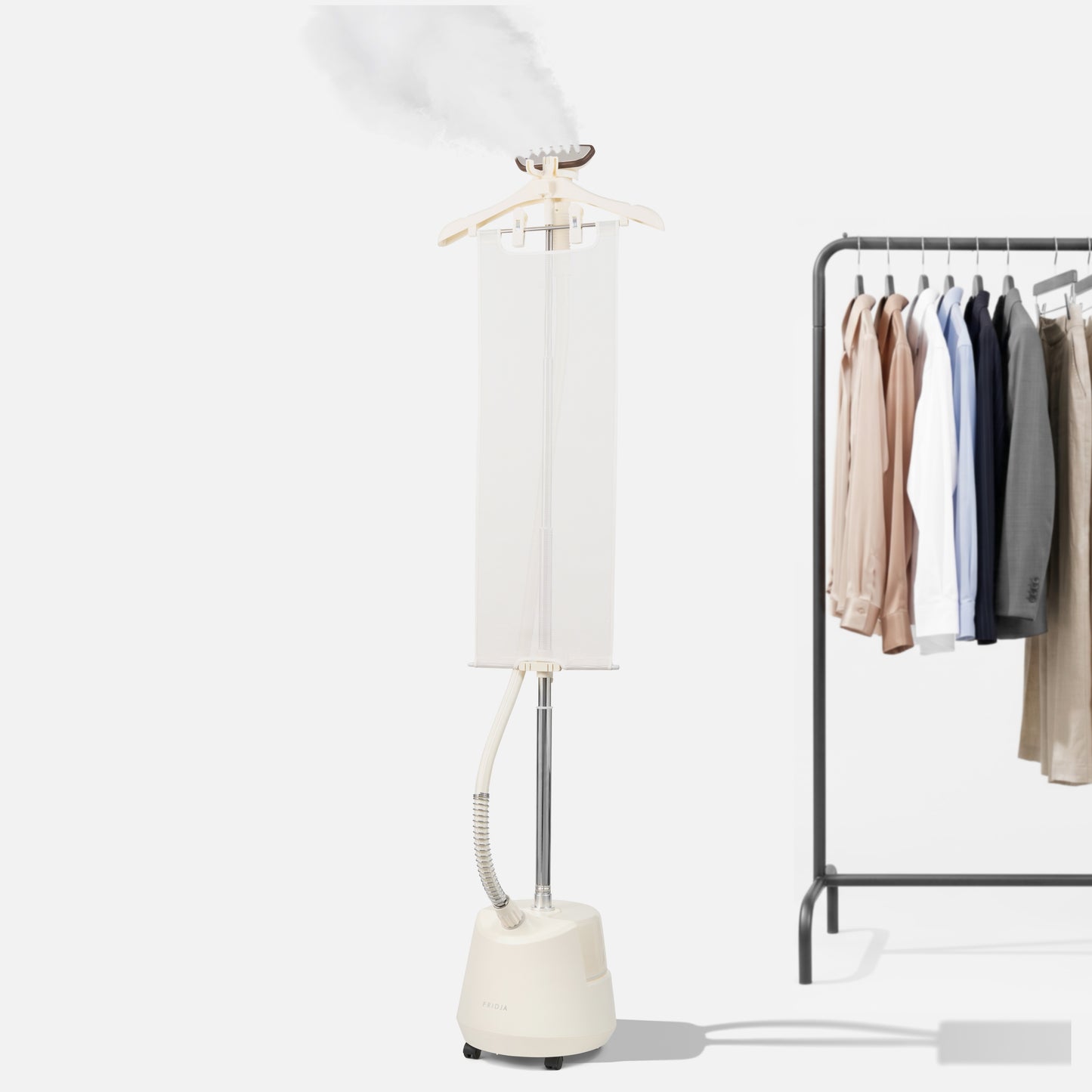 The Fridja f1200 Clothes Steamer Pro releases steam next to a clothing rack with neatly hung shirts, jackets, and pants against a plain white background, highlighting the effortless wrinkle-removing power of this advanced garment steamer.