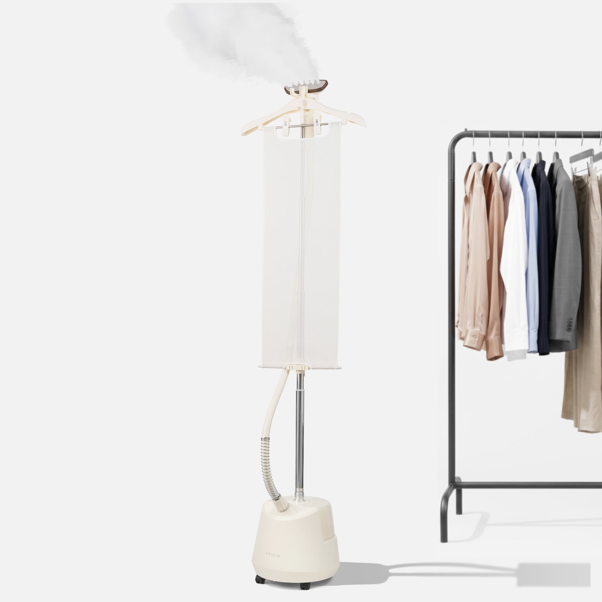 The Fridja f1200 Clothes Steamer Pro releases steam next to a clothing rack with neatly hung shirts, jackets, and pants against a plain white background, highlighting the effortless wrinkle-removing power of this advanced garment steamer.