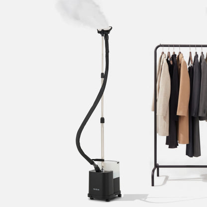 The Fridja f1600 Commercial Clothes Steamer emits steam next to a clothing rack filled with various jackets and coats, highlighting professional garment care against a plain white background.