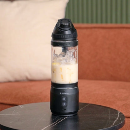 The Fridja f500 Portable Blender, ideal for making a protein shake or smoothie wherever you are, is pictured filled with iced coffee on a round black marble table set in front of a rust-colored couch and green-striped wall.