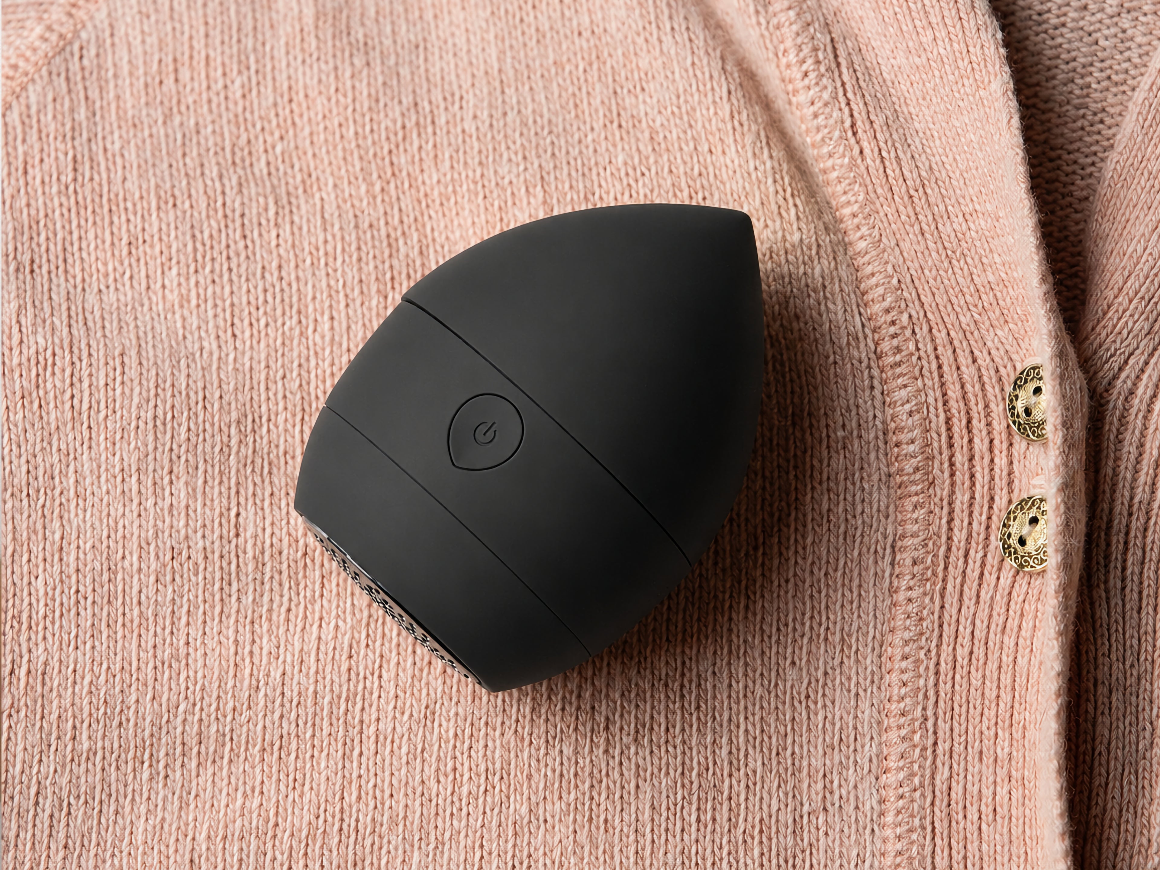 A black, egg-shaped electronic device with a power button symbol sits on a pink textured fabric with gold decorative buttons.