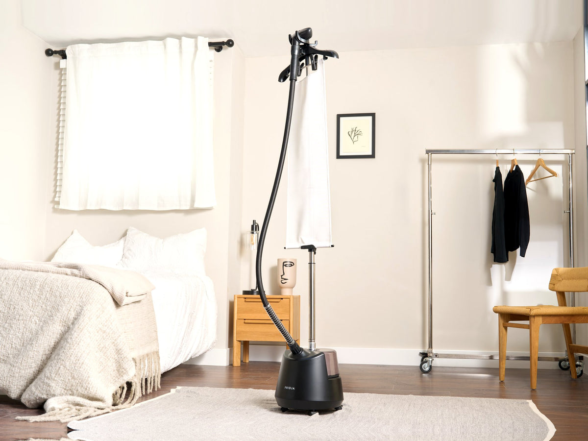 Fridja f1200 Clothes Steamer Pro standing in a bright bedroom beside white curtains