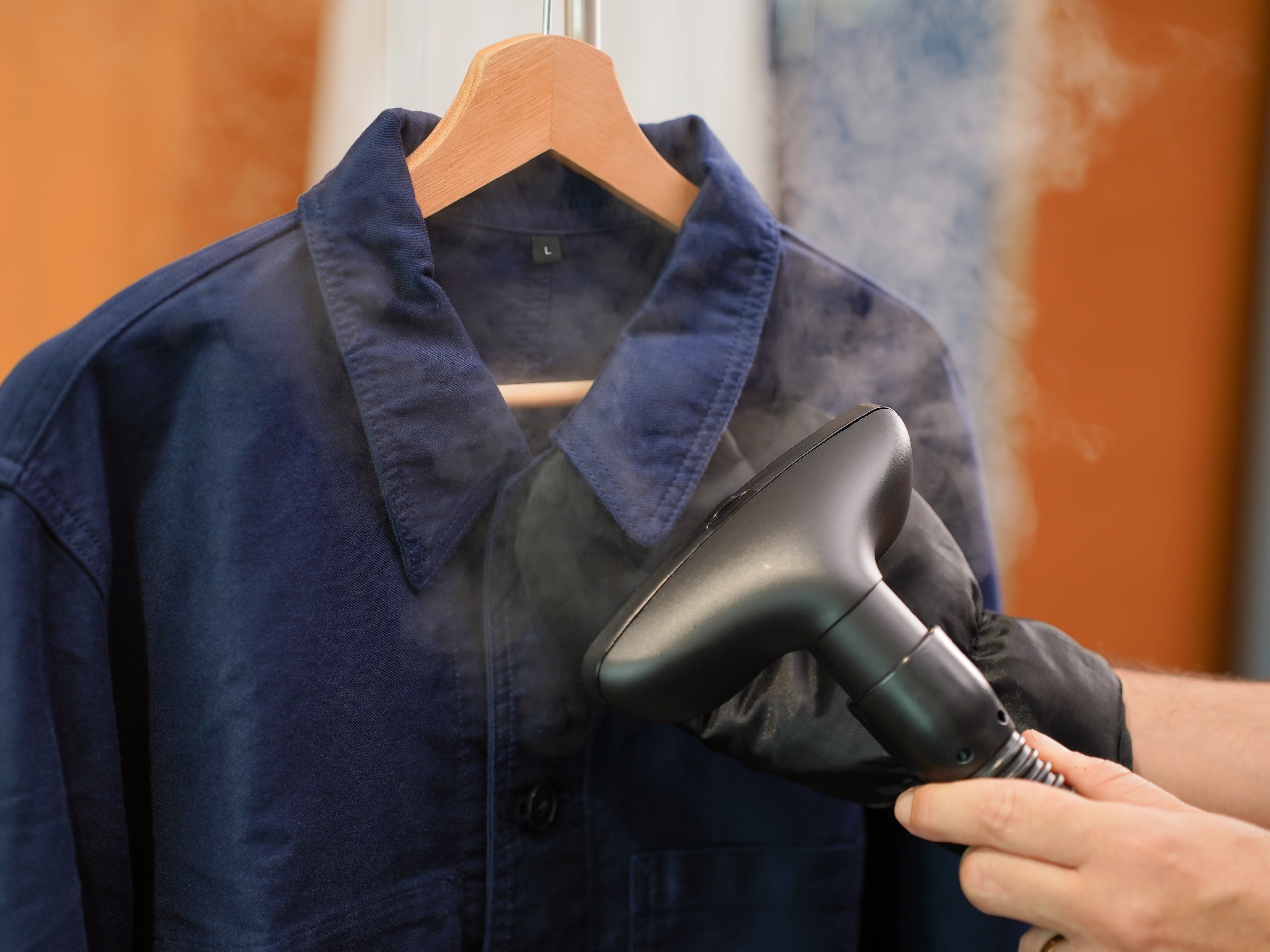 A person uses a handheld steamer to remove wrinkles from a blue button-up shirt hanging on a wooden hanger, with steam visibly rising from the fabric.