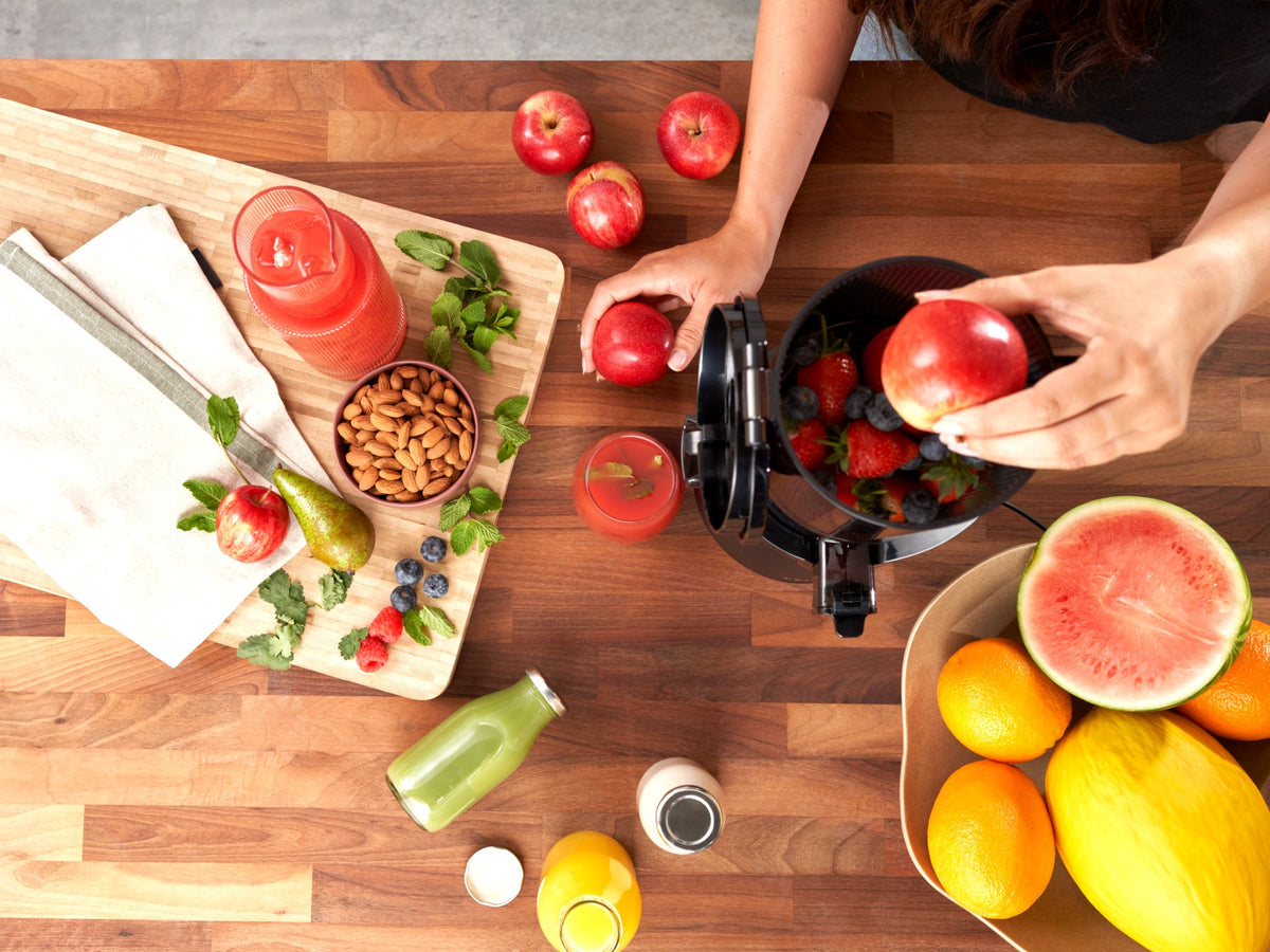 Overhead view of the Fridja f2500 Self Feeding Whole Fruit Juicer being loaded with apples on a wooden worktop surrounded by fresh fruit and juices
