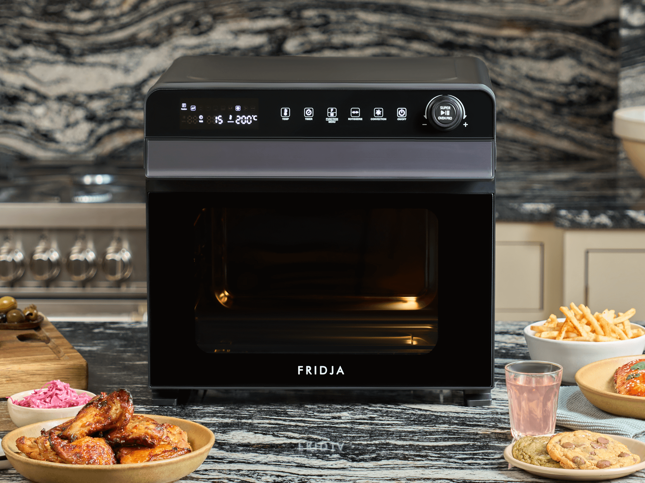 A black Fridja countertop oven sits on a marble kitchen counter, surrounded by plates of cooked chicken wings, fries, cookies, a glass of pink drink, and a bowl of slaw. The oven display is lit up with settings.