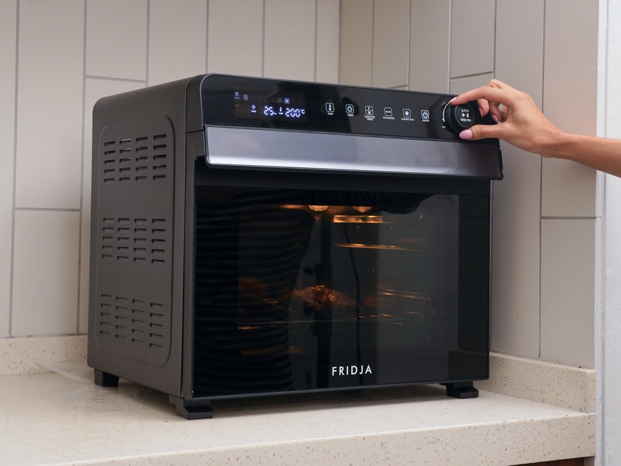 A person adjusts the settings on a black Fridja countertop oven with an illuminated digital display, positioned on a beige kitchen counter against a tiled wall.