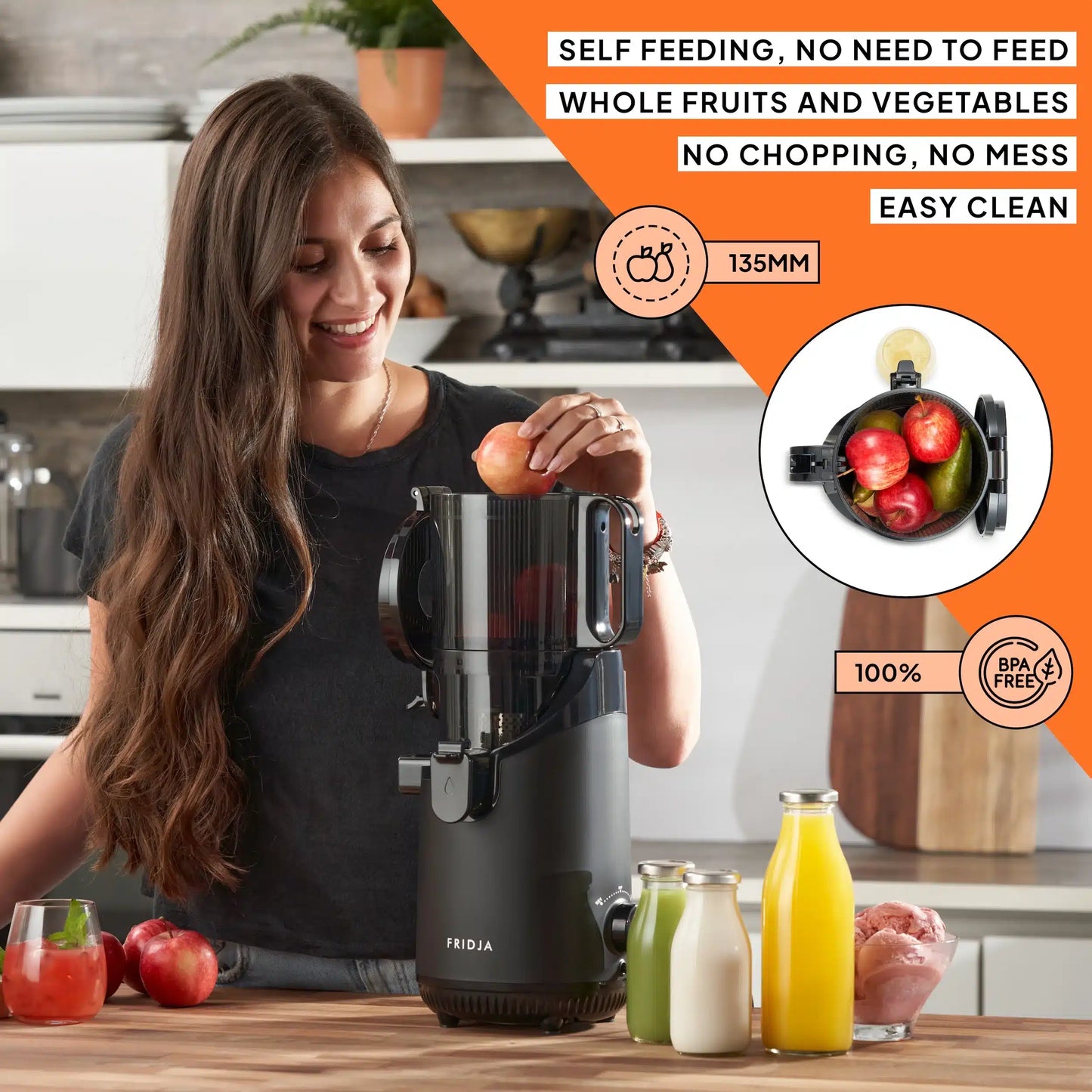 A woman uses the Fridja f2500 Self Feeding Whole Fruit Juicer, placing an apple into the black appliance on a kitchen counter with apples, oranges, juice bottles, and chopped fruit. Features include cold-pressed juice, BPA-free build, self-feeding, and easy cleaning.