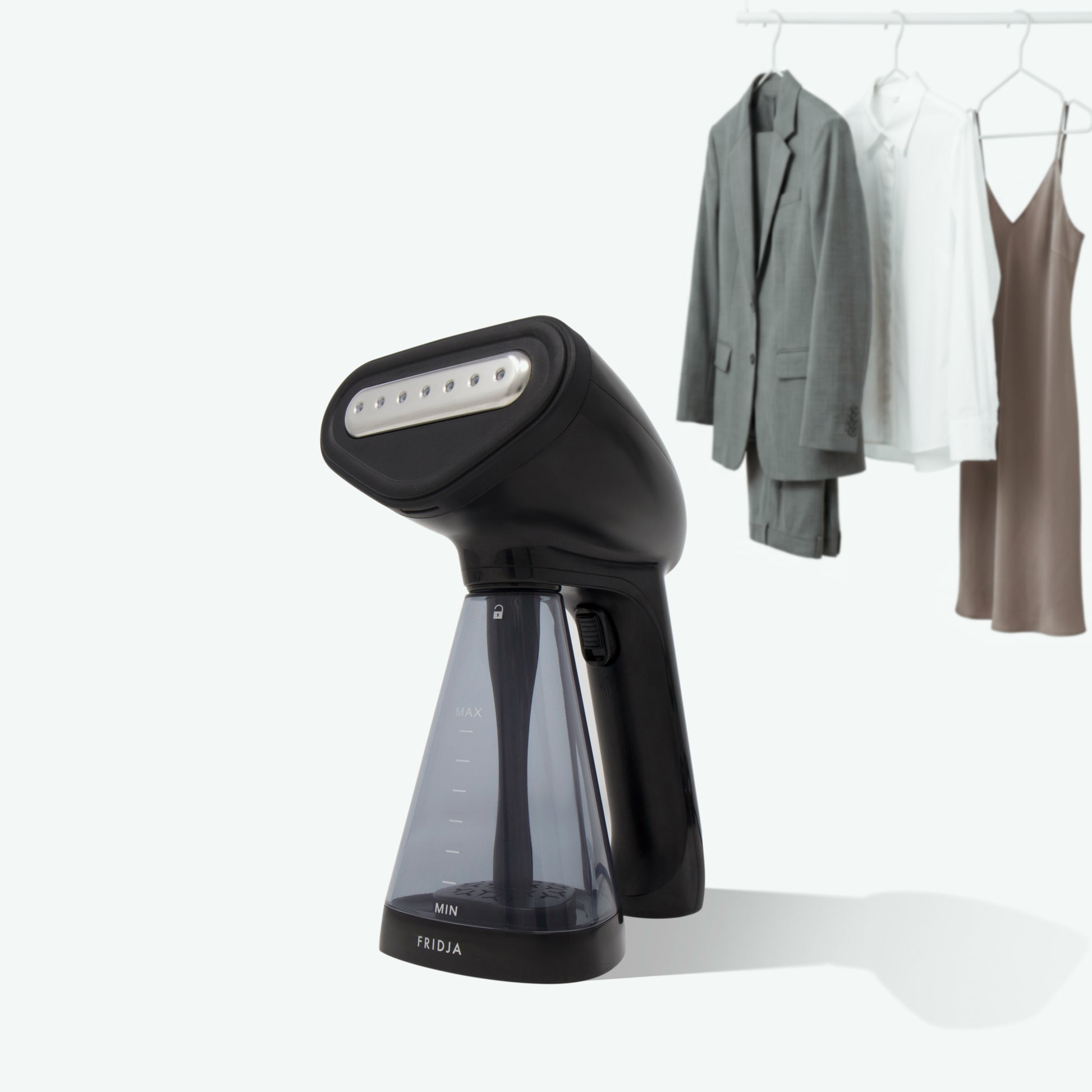 A Fridja f10 Handheld Clothes Steamer with a transparent water tank sits in the foreground; in the background, a gray suit, white shirt, and brown dress hang on a rack.