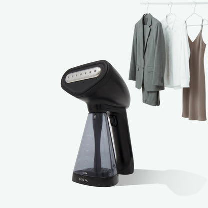 A Fridja f10 Handheld Clothes Steamer with a transparent water tank sits in the foreground; in the background, a gray suit, white shirt, and brown dress hang on a rack.