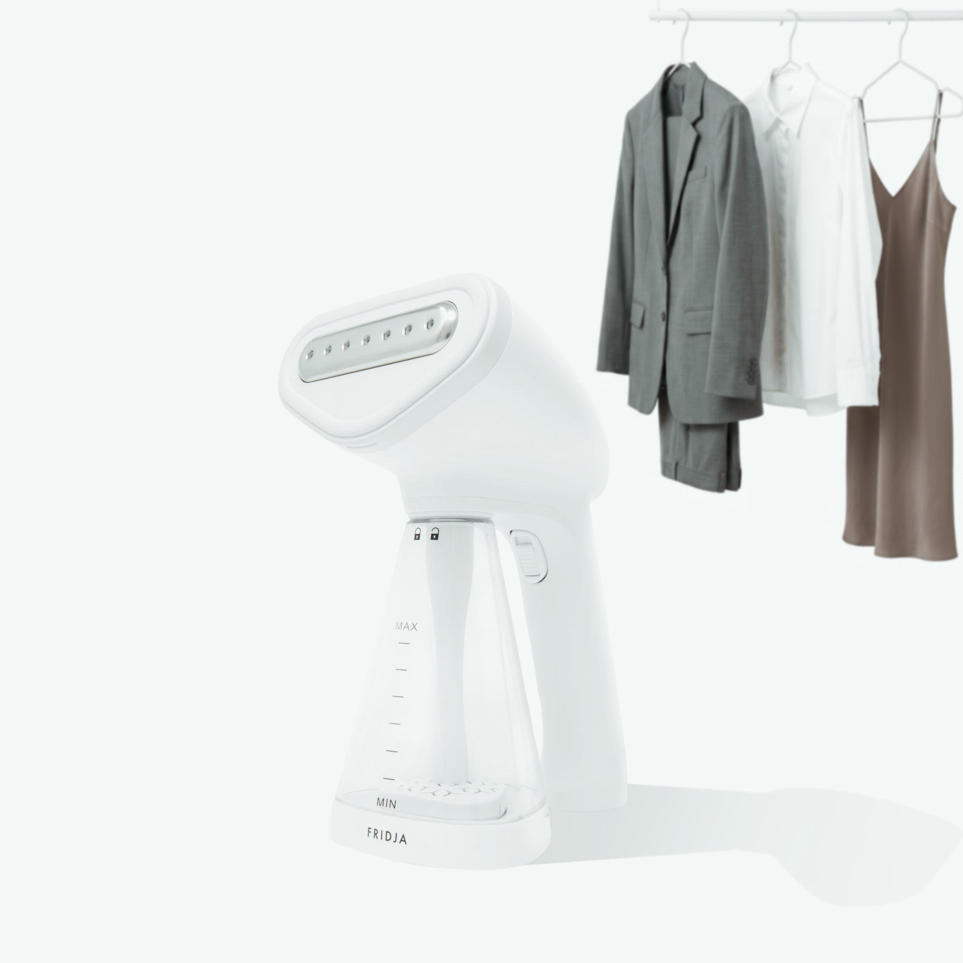 The Fridja f10 Handheld Clothes Steamer is featured in the foreground against a minimalistic white backdrop, highlighting its sleek design and practical functionality. In the background, a gray suit, white shirt, and brown dress hang neatly on a clothes rack.