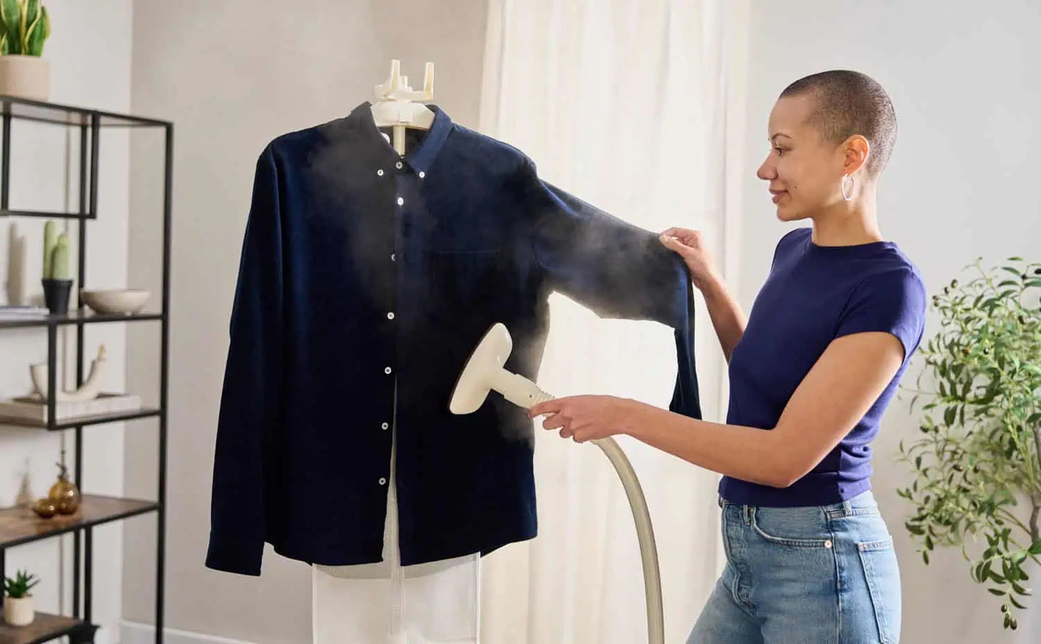 A person uses a handheld garment steamer to remove wrinkles from a dark button-up shirt hanging on a stand in a bright, modern room with plants and shelves in the background.