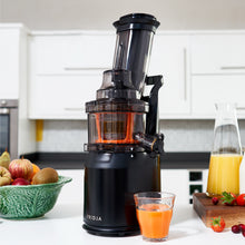 A  Fridja f1900 Whole Fruit Juicer with Cold Press Technology sits on a kitchen counter filled with orange juice, surrounded by a glass and carafe of juice, fresh fruit in a green bowl, and strawberries on a cutting board.