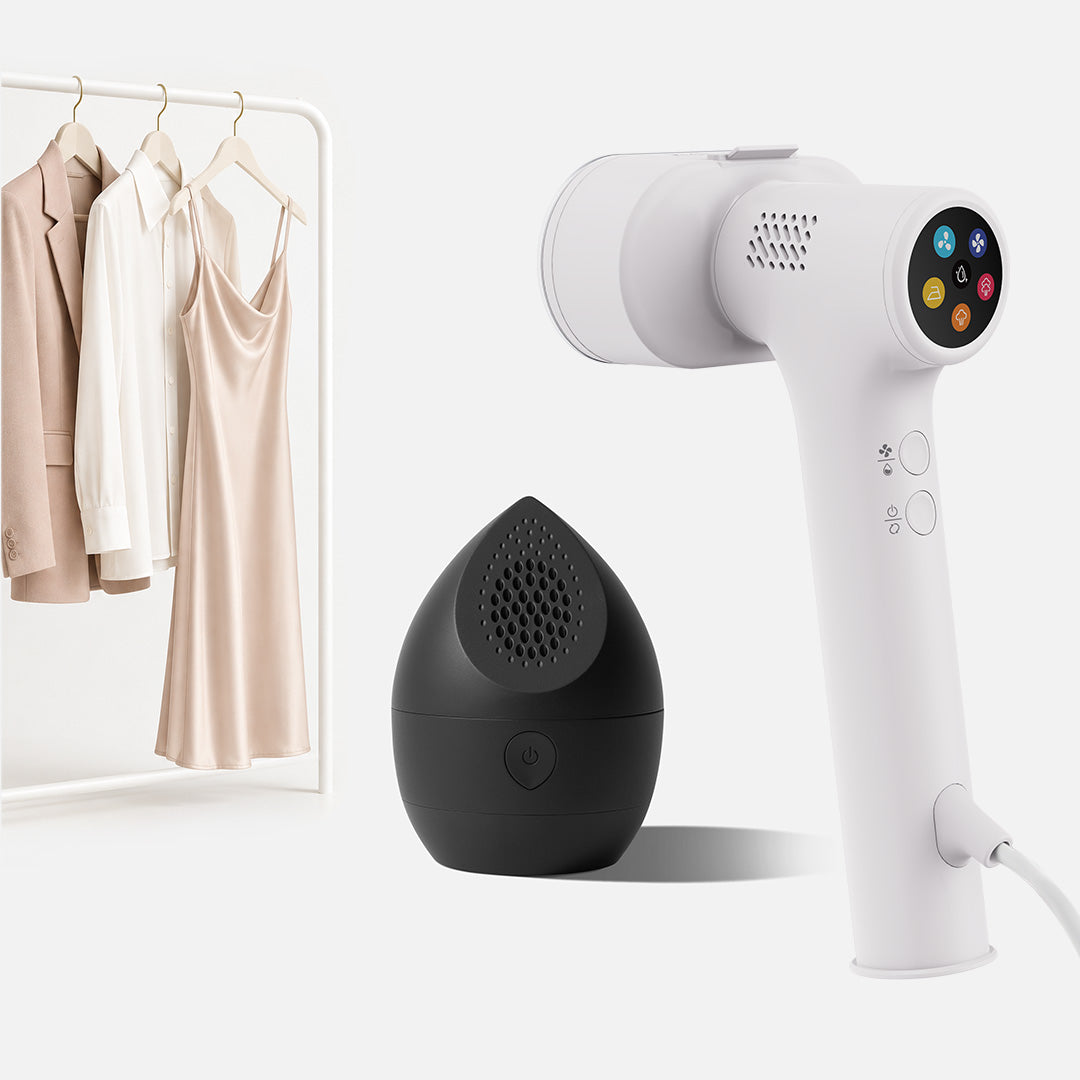 A white Fridja Handheld Garment Care Pro Duo—featuring the f20 Handheld Clothes Steamer Pro with control buttons—stands beside a sleek black, teardrop-shaped f40 Fabric Shaver. In the background, three light-colored garments hang neatly on a rack, creating a minimal and clean scene that highlights effective garment care.