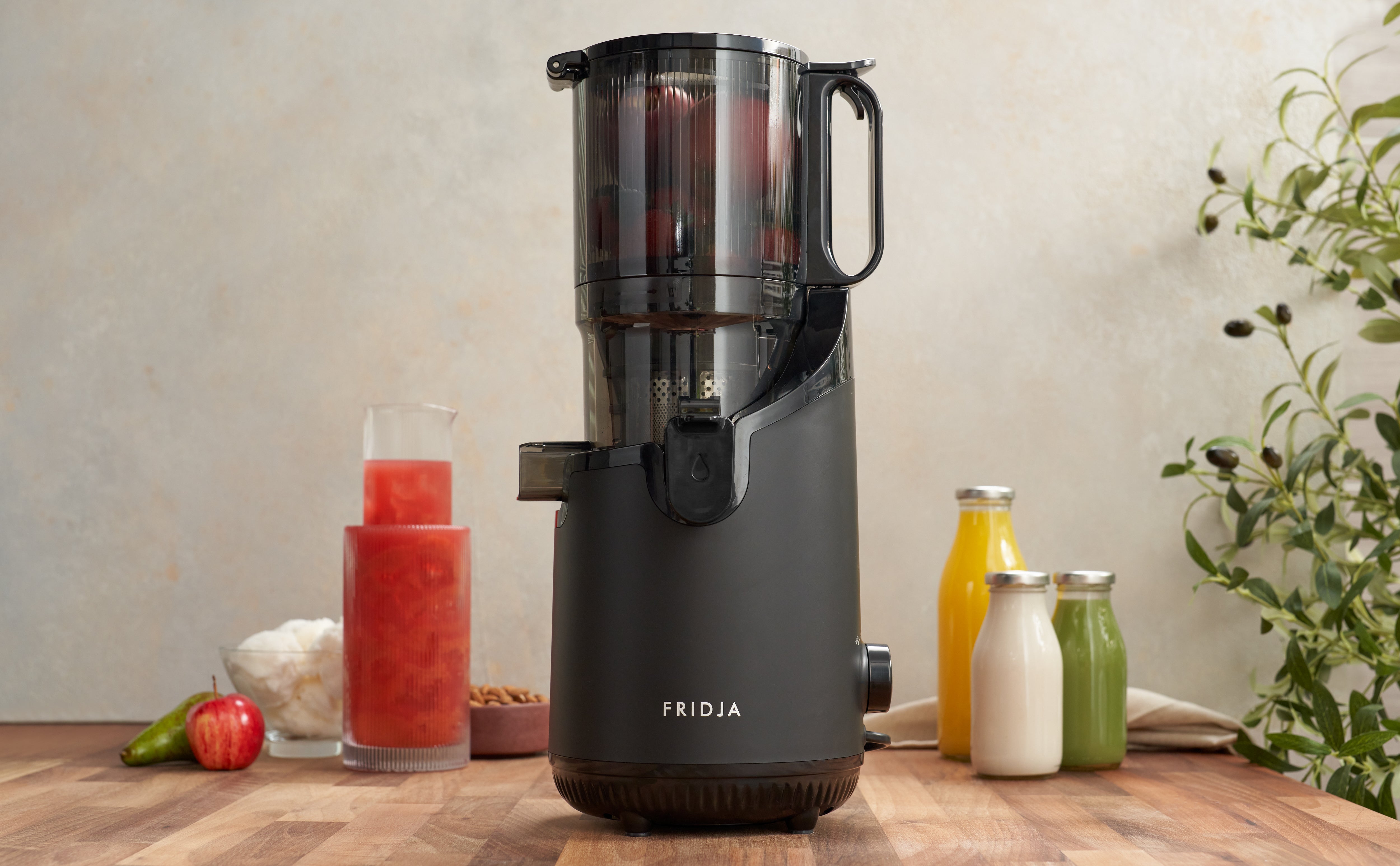 A sleek black juicer labeled “FRIDJA” sits on a wooden counter with jars of fresh juice, an apple, and ingredients nearby. There’s a plant and a light-colored wall in the background.