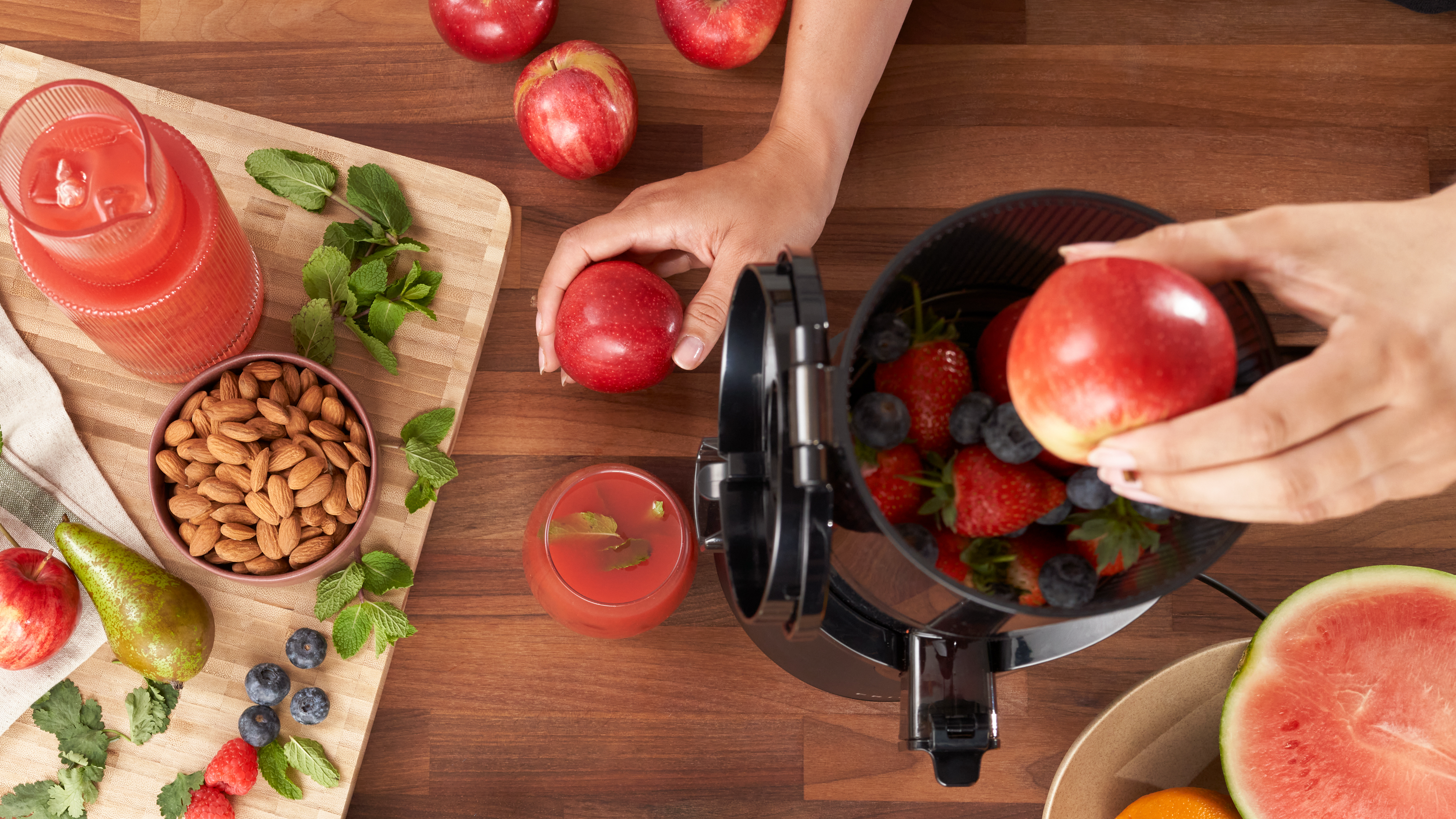A person places an apple into a juicer filled with strawberries and blueberries. Nearby are apples, almonds, mint, a pear, watermelon, and glasses of juice on a wooden table.