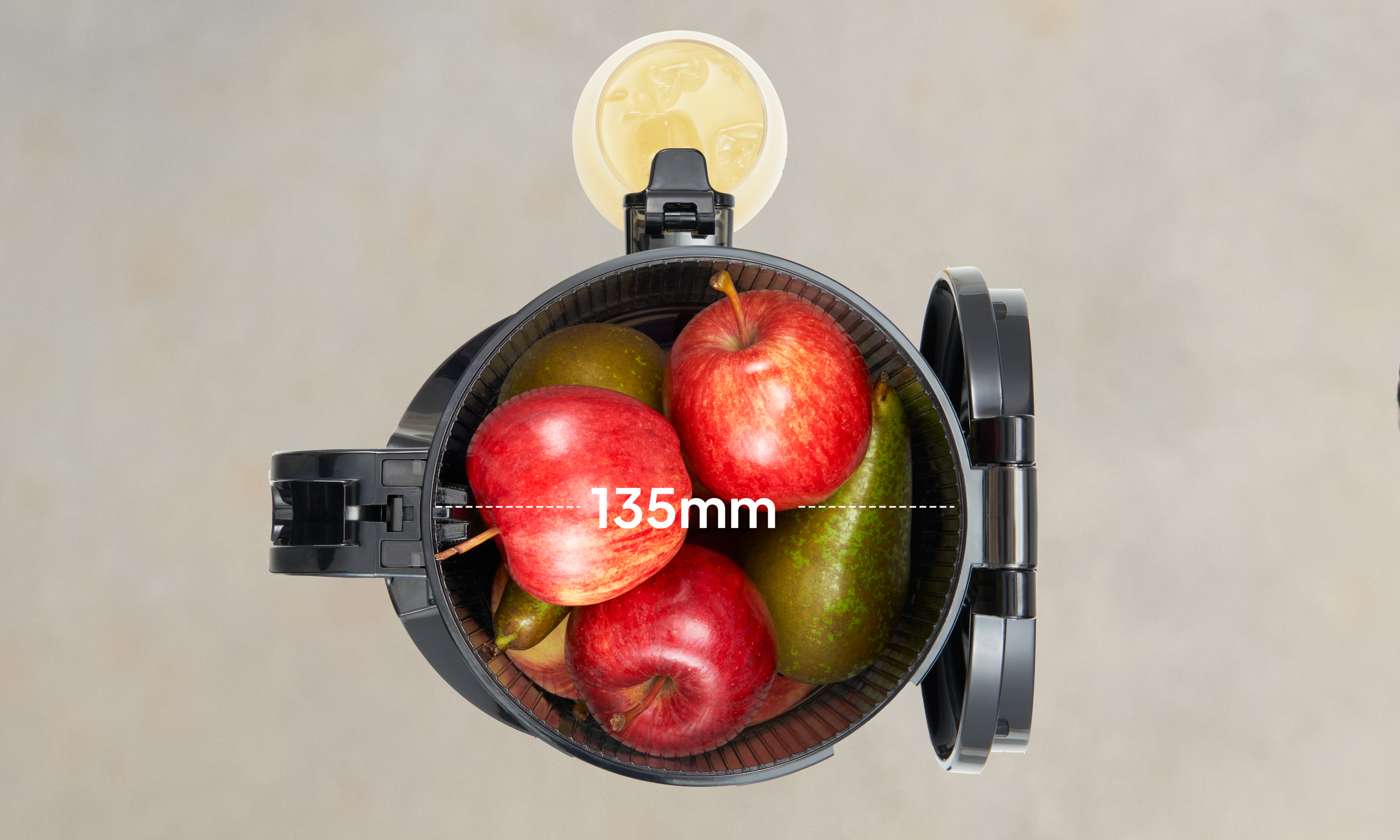 Top view of an open air fryer basket filled with apples and pears, with a dotted line labeled 135mm showing the internal diameter. A small cup of yellow liquid is placed above the basket.