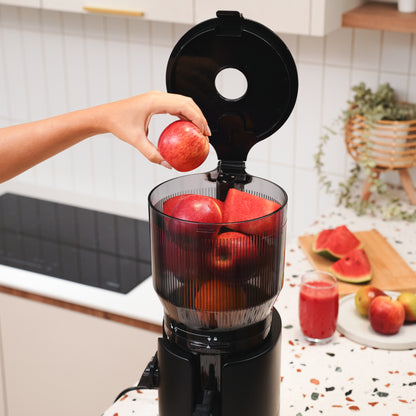 A person places a red apple into a blender filled with more apples. The kitchen counter in the background has a cutting board with watermelon slices and a glass of red juice.