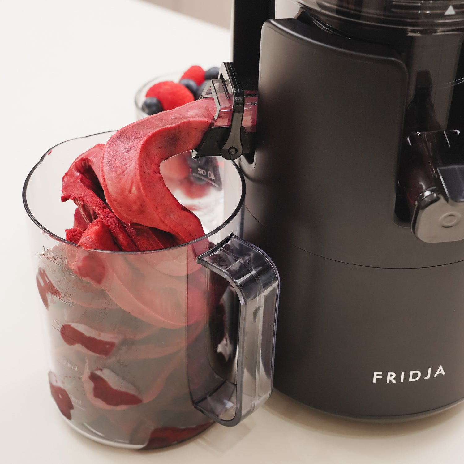 A close-up of a black Fridja machine dispensing thick, red berry sorbet into a clear container, with a bowl of mixed berries blurred in the background.