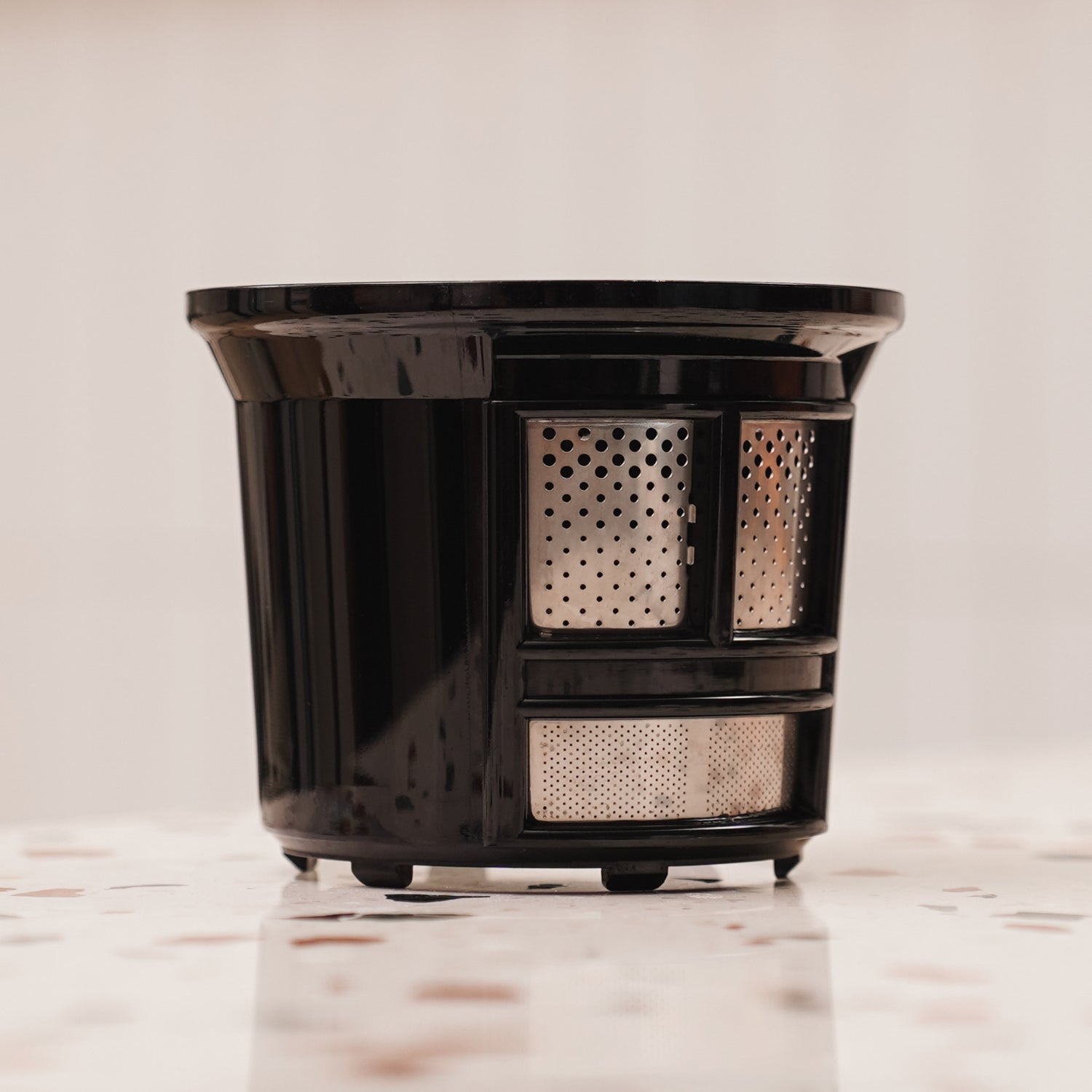 A black cylindrical tea or coffee filter with fine metal mesh panels, standing upright on a speckled white surface, against a light blurred background.