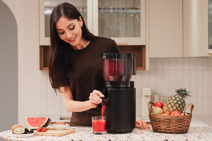 A woman in a brown shirt smiles as she pours fresh red juice from a black juicer into a glass. A basket of fruits, including a pineapple and apples, sits on the counter beside her in a modern kitchen.