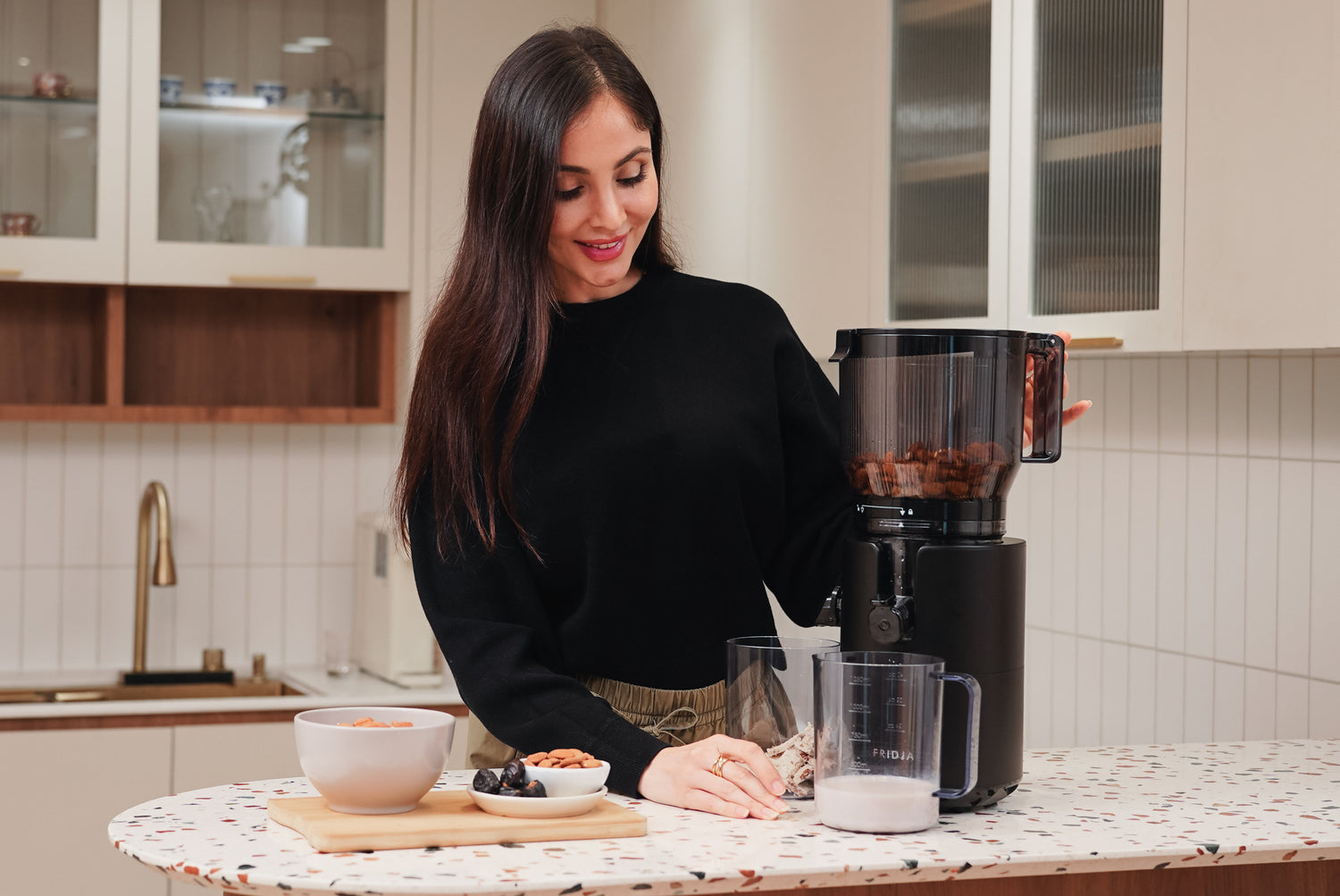 A woman with long brown hair stands in a kitchen using an electric grinder or food processor to grind nuts, with a bowl of whole nuts and a plate of dates and chocolate nearby on a countertop.