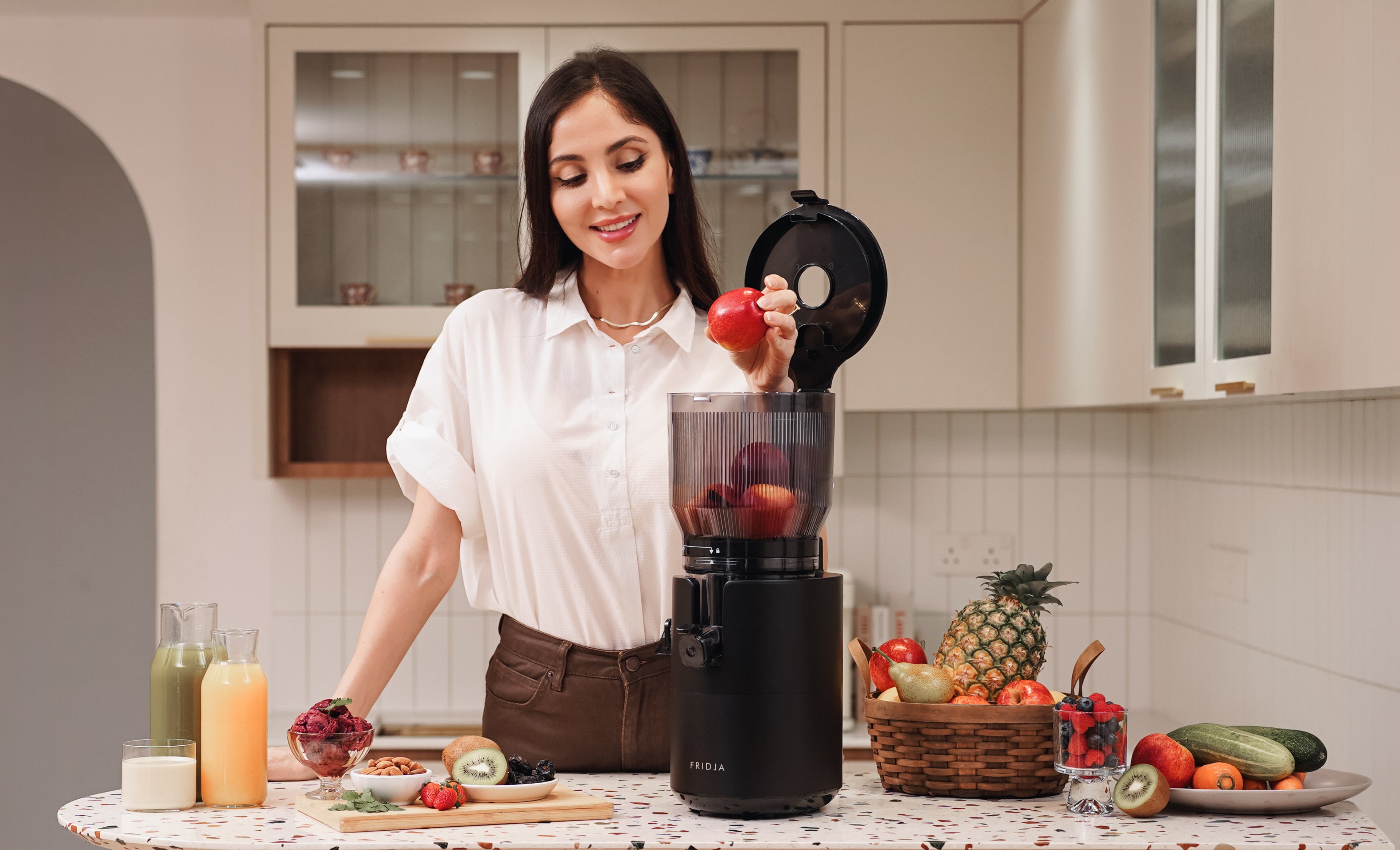 A woman in a white shirt smiles while placing apples into a black juicer on a kitchen counter, surrounded by various fruits and glasses of juice. The kitchen is bright with light-colored cabinets.