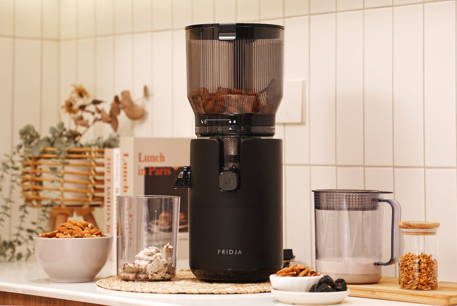 A black Fridja nut milk maker sits on a kitchen counter. Surrounding it are bowls of nuts, a clear tumbler with nut pieces, a jug of nut milk, and kitchen decor including a book and a plant.