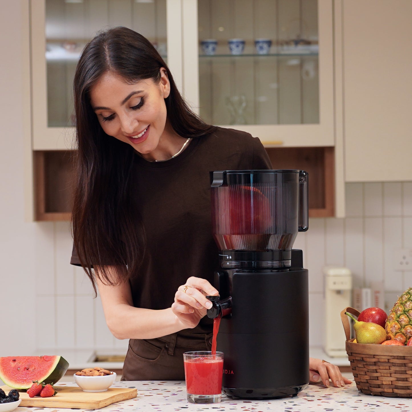 A woman in a brown shirt smiles while pouring fresh watermelon juice from the Fridja f2800 Open Whole Fruit Juicer into a glass in her kitchen, with sliced fruit and a basket of pineapple on the counter beside her.