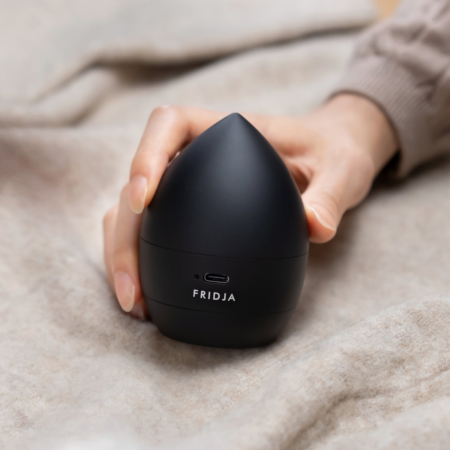 A hand holds a sleek, black Fridja f40 Fabric Shaver with a USB-C port, resting on beige fabric—ideal for keeping clothes lint-free and fresh.